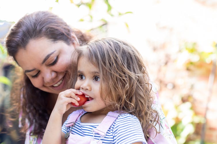 A mother holds her young daughter while the child eats a tomato