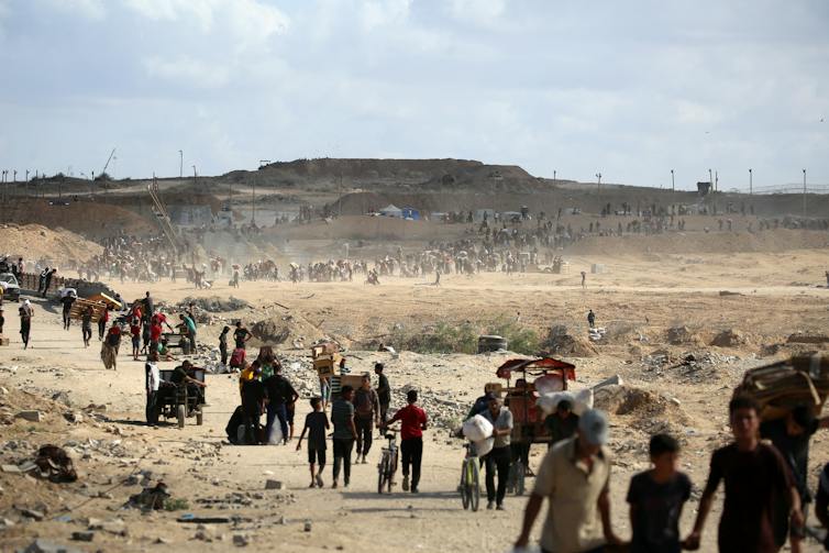 A long line of people with empty cardboard boxes and plastic bags walk through an arid landscape.