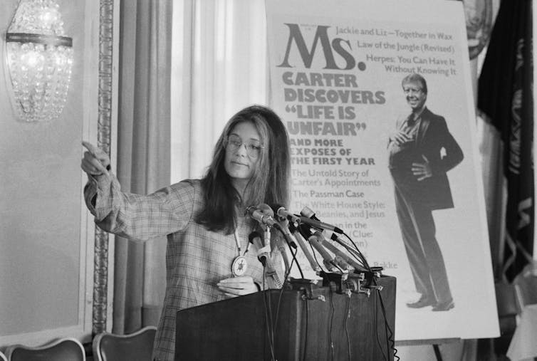 A woman stands at a lectern in front of a large magazine cover and speaks.