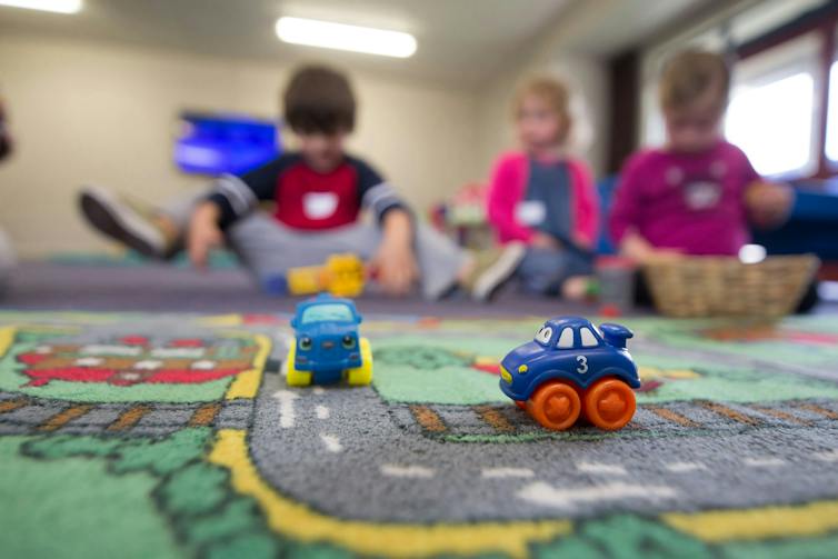 Some toy cars on a floor rug with small children in the background