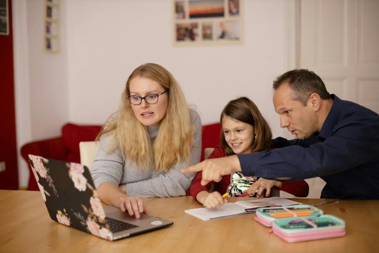 Parents sit at a table either side of their daughter to help her study
